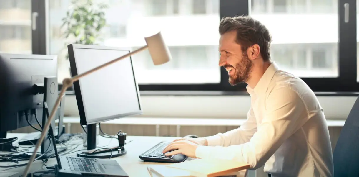 A man smiling while working at an office desk with a computer and natural daylight streaming in through large windows.