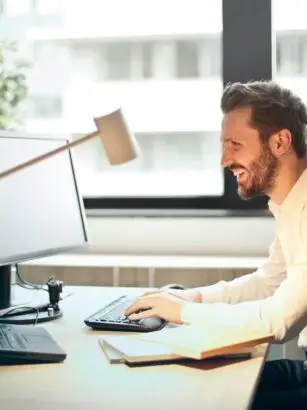 A man smiling while working at an office desk with a computer and natural daylight streaming in through large windows.
